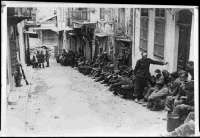 German POWs at Canea, Crete, 1941. Shows a row of German paratroopers under New Zealand guard lined up along one side of a street. Some are wounded.