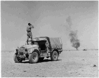 New Zealand soldier Arch Currie standing on a Morris truck at El Alamein, Egypt, looking though binoculars. Original caption reads: 'NZ Div hold off enemy for whole day covering withdrawal of other troops to El Alamein. Shows one of our trucks burning.' Photograph taken circa 9 July 1942 by H Paton.