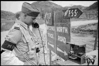 A New Zealand Division Provost (J J Morgan of Pukekohe) communicating on the telephone with another traffic post, at the commencement of the Inferno Track, in the Cassino area, Italy. Shows Morgan talking into the phone receiver, beside a sign that points in one direction to the 