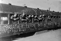 Soldiers leaning out of a train as they depart to serve in World War II.  The train is covered in graffiti including New Zealand place names and phrases such as 